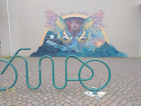 I'm the foreground, a green metal bent pipe bike rack in the shape of a stylized bicycle. I'm the background a mural on a tan wall, consisting of a mountain range in dark cool colors, a pair of disembodied wings above them in warm colors, with a text banner over an arch reading "Alis volat propriis."