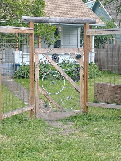 A wood framed gateway to a grassy yard. There is a bell hanging from the middle of the gateways top beam, and the space in the gate itself is filled by old bicycle wheels of various sizes, as well as chainrings, cranks and cogs 