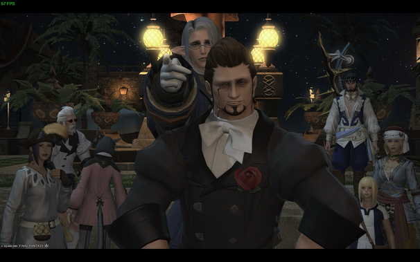 A group of Final Fantasy XIV (FFXIV) player characters are gathered at night. In the front stands a large, imposing male character with dark hair and a trimmed goatee, wearing a black tuxedo adorned with a red rose boutonniere. He has a monocle and a slight smirk. Behind him, slightly to the left, is a white-haired male character wearing glasses, pointing his fingers like a gun toward the camera, and several other characters are arranged around them. The scene is set in what appears to be an outdoor area with palm trees and decorative lighting. The background is a night sky.