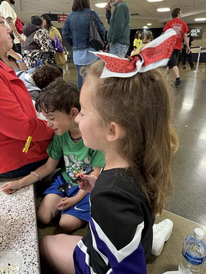 A girl wearing a cheerleading outfit with a large red and white bow sits next to a boy in a green shirt. They are in a crowded indoor space, with adults and children milling about in the background. The girl is holding a spoon 