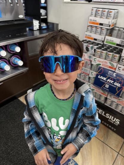 A young boy is smiling while wearing oversized blue sunglasses. He is dressed in a green shirt with cartoon characters and a plaid jacket. The background shows beverage dispensers and shelves stocked with energy drinks.