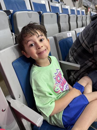 A young boy with wet hair is sitting in a stadium seat, wearing a bright green shirt and blue shorts. He has a cheerful expression and is posed slightly sideways, with a few empty seats visible in the background.