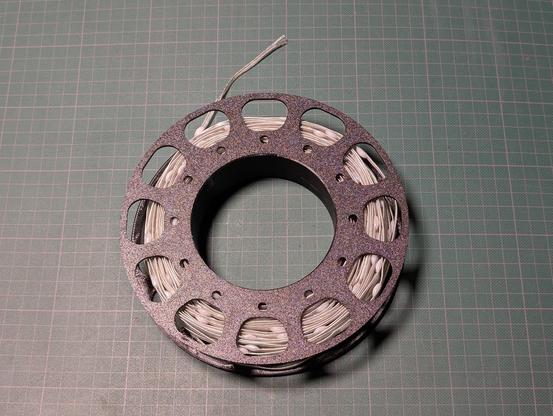A black 3D printed spool containing a 40 meters long strand of white addressable pebble LEDs. It's sitting on a green grid cutting mat as a backdrop.