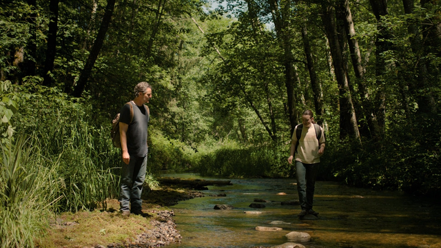 Cena da série The Last of Us mostra os personagens Joel e Ellie caminhando por uma floresta durante o dia. Joel (Pedro Pascal), à esquerda, está parado na margem de um riacho, vestindo camiseta escura, jeans e mochila. Ellie (Bella Ramsey), à direita, atravessa o riacho pulando entre pedras, usando uma camiseta clara, jeans escuro e mochila. Ambos estão cercados por árvores altas e vegetação densa, com luz solar filtrando pelas folhas, criando um clima tranquilo e natural.