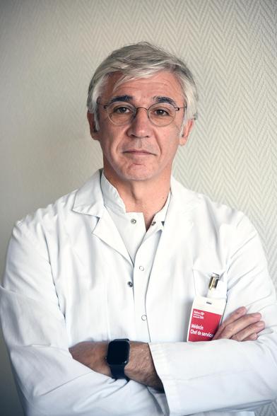 Portrait du Pr Jean-Claude Deharo en blouse blanche, bras croisés, portant une étiquette “Médecin – Chef de service”, debout devant un mur clair à motif discret.