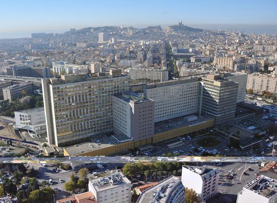 Vue aérienne de l’Hôpital de La Timone à Marseille, avec ses grands bâtiments hospitaliers, en arrière-plan le centre-ville et la basilique Notre-Dame de la Garde en hauteur.