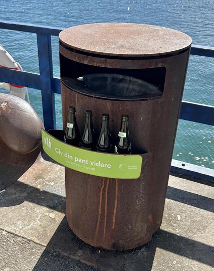 A cylindrical trash bin located near a body of water, designed for collecting bottles. It has a green sign in Danish that translates to "Donate your deposit," indicating that users can return their bottle deposits here. Several glass bottles are displayed in a holder.
