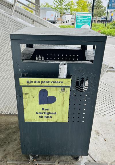 A black trash bin with a yellow sign featuring a blue heart, promoting cleanliness in Copenhagen. A partially visible can is placed in the bin. The background includes a staircase and trees.