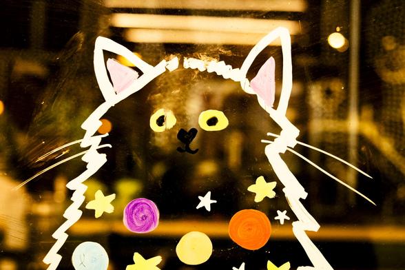 On shop window glass, pictured from the street at night, a cat is drawn with thick coloured marker. The animal has bright yellow eyes and appears to be smiling in a quizzical way at passers by. Its body is transparent and decorated with stars and circles