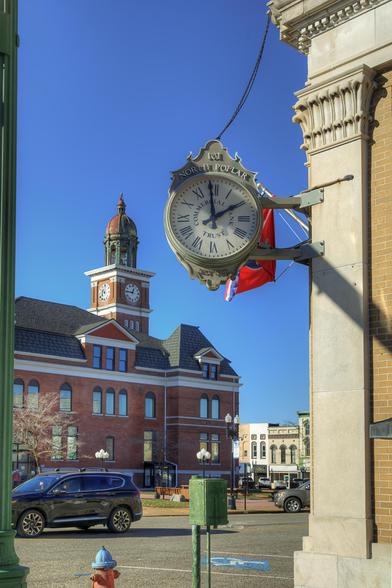 A large clock hangs on the side of a building, set against the backdrop of a historic red brick courthouse with a clock tower.