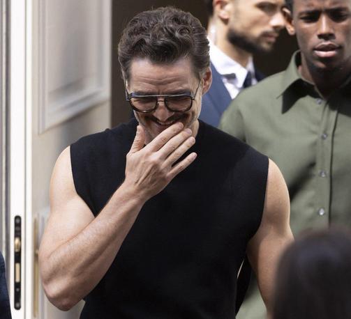Pedro Pascal in Cannes. He wears all black trousers, shoes and a muscle shirt without arms that show off his biceps. He smiles and scratches his chin.