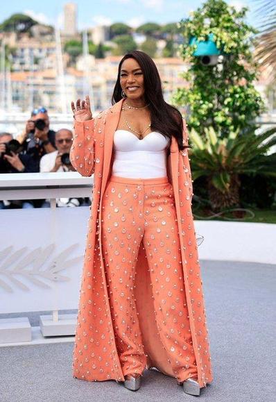 Angela Bassett in a salmon suit with a coat and white bustier. The suit has silver embelishments on it that look like hanging teardrops. She wears long open black hair and silver high heels.