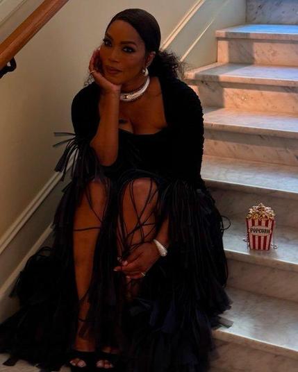 Angela Bassett sitting on stairs after a premiere in Cannes. She wears a black form-fitting dress with  bolero jacket with long chiffon trumpet sleeves. She wears her long black hair in a curly ponytail and silver jewelry. Next to her stands a cute handbag that looks like a pocorn box with red and white stripes and popcorn in it.