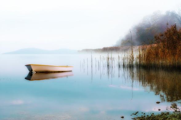 A lone boat floats gently on the surface of a calm lake, its pale yellow hull reflecting subtly in the still water. A soft mist drapes over the landscape, blending the distant hills into the sky, creating an ethereal, almost dreamlike atmosphere. Tall, golden reeds emerge from the water’s edge, their delicate stems reaching skyward, mirrored in the glassy lake below. On the right, a wooded shoreline shrouded in fog adds a sense of quiet mystery, its muted autumn colors barely visible through the haze. The entire scene exudes tranquility, solitude, and a deep connection to nature—a fleeting moment of peace suspended in time.
