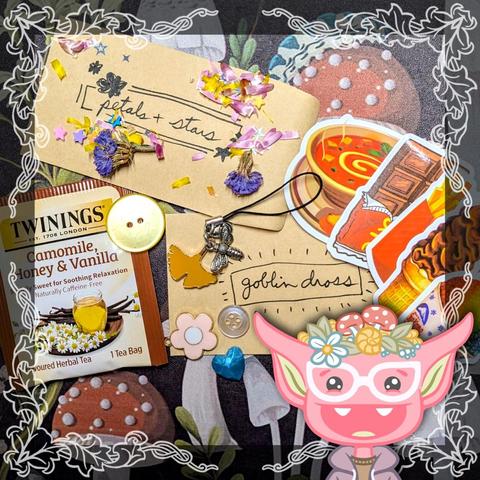 Photo of the contents of a Goblin Dross package, all ready to be put in an envelope and mailed out. It includes a Camomile, honey and vanilla tea bag, two random buttons, a metal flower charm, various food item stickers, a blue resin heart, and a handmade luck dangle with a silver bee and yellow ginkgo leaf charms attached. A small envelope hand-labelled "goblin dross" sits in the middle of all of this month's items. And a small envelope hand-labelled "petals and stars" sits on top, covered in flower petals and star confetti. At the bottom right of the image is an illustration of a smiling pink goblin with white glasses and a purple hoodie, wearing flowers and mushrooms and a snail shell on her head.