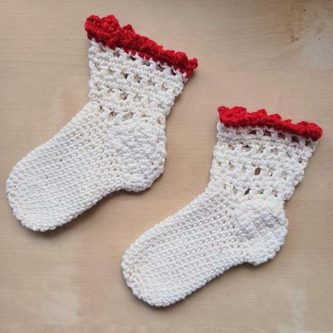 Flat lay view of 2 white crochet baby socks with a lace edging and red picot finishing on a light wooden background.