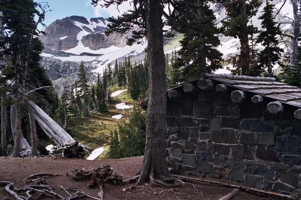 Set among conifer trees is a stone masonry trail shelter with a shingled wood-beam roof, with a steep mountain wall rising in the background, partially covered by snow.