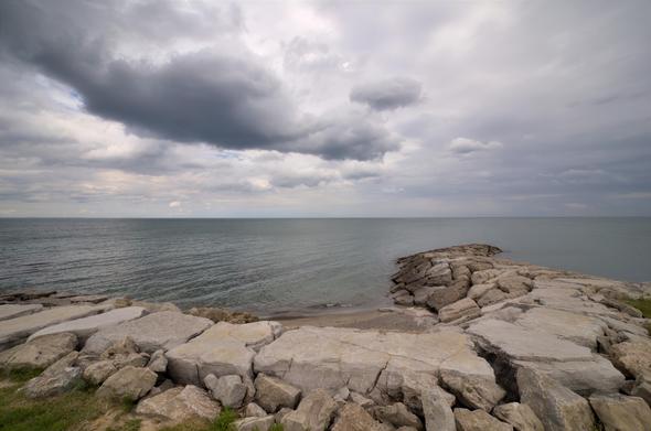 From a slightly elevated but broad-angle view, a curved breakwater of massive, roughly hewn limestone slabs fills the lower half of the frame. The blocks, varying in size and punctuated by cracks and flecks of pale yellow lichen, extend into the lake as a narrow jetty. Beyond, the water’s surface shows fine, uniform ripples. Above, thick layers of cloud—ranging from light dove-gray to deeper charcoal—stretch across the sky, diffusing the daylight into even, shadowless illumination.