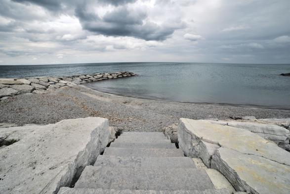 Seen from directly above a short flight of wide stone steps, this image guides the eye down the worn treads to a narrow gravel-and-sand shoreline. On either side of the steps, flat limestone platforms show faint scratches and weather stains. Just past the beach, another stubby breakwater of stacked boulders reaches into the lake. The water here is glassy close to shore, darkening to steel blue at the horizon. Overhead, heavy cloud cover creates a hushed, almost monochrome scene that feels both peaceful and vast.