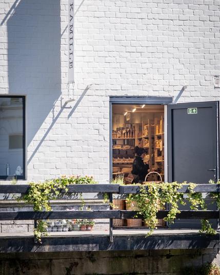 A white brick building with an open dark doorway with an exit sign reveals a glimpse of a small shop interior with shelves of products and a person inside. The building sits alongside what appears to be a canal, with a black railing adorned with climbing plants and vines. Baskets and potted plants rest on the dock-like platform in bright sunlight, creating strong shadows against the clean white wall.