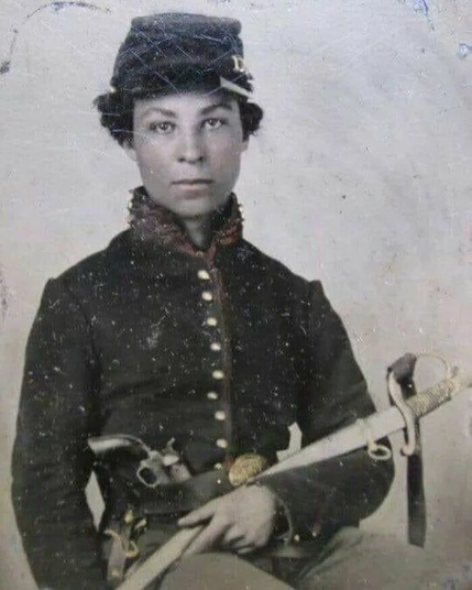 The image depicts a person dressed in a historical military uniform, likely from the American Civil War era. The individual is wearing a dark, high-collared jacket with gold buttons down the front, and a matching dark hat with a small brim. The uniform is complemented by a belt with a large, ornate buckle, and the person is holding a sword with a white hilt and a curved blade, suggesting a cavalry or infantry role. A revolver is visible in a holster on the belt. The background is plain and light-colored, emphasizing the subject. The photograph has a vintage appearance, with visible signs of aging and wear, including scratches and discoloration.