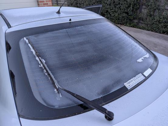 Photo of the rear windscreen of a silver coloured sedan. The glass is frosted over and the windscreen wiper has managed to push only some of the ice off to the side. 