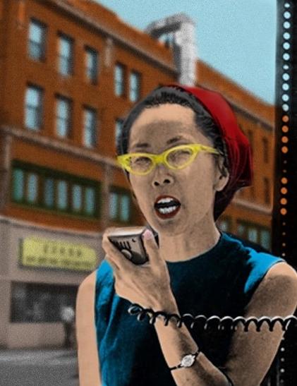 Yuri Kochiyama with glasses and a red headscarf is speaking into a vintage megaphone mic after Malcolm X’s death. She is wearing a blue top and appears animated. The background features a cityscape with brick buildings.