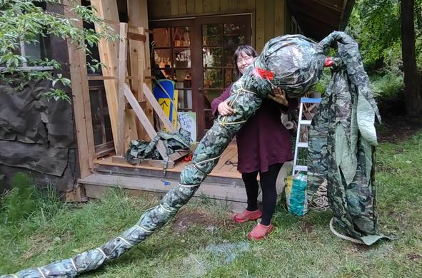 Artist Emily van Lidth de Jeude stands smiling on the lawn in front of her home-studio holding a giant bull-kelp sculpture made of green camouflage tarp and red Tuck tape. The head of the bull-kelp is about two feet in diameter, and the leaves hanging off the end of it are seven feet long. A very long eight-inch-wide stem goes off to the left and out of the photo. In the background can be seen the wooden porch of Emily's studio, with an easel and some boards stacked on it, some of the tarp remnants from creating the sculpture, and a small metal step-ladder. Emily is wearing red shoes, black leggings, and a very long burgundy sweater.