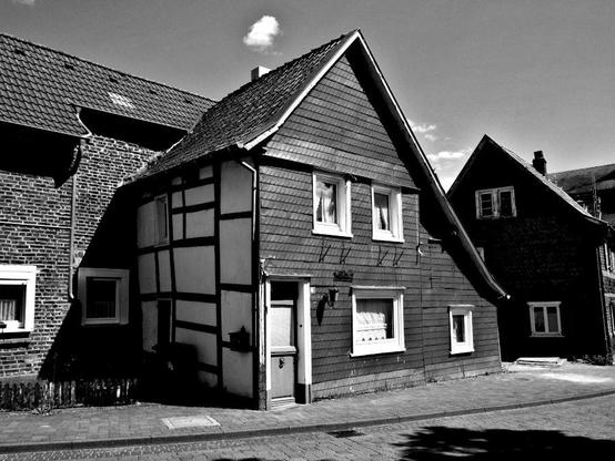 A house typical of Germany's Bergisches Land. One side has characteristic slate siding, another has visible timber framing warped with age. The doors and windows have white molding. The roof is steeply pitched. It is probably 200 or 300 years old.