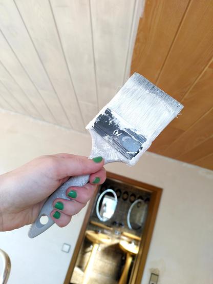 Fee hält einen Pinsel voller weißer Farbe hoch, im Hintergrund eine Holzdecke, halb grundiert. Schemenhaft durch eine Tür zu erkennen ein Siebzigerjahre-Bad.