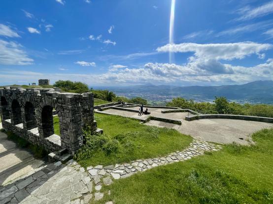 A scenic view from a hillside with stone ruins and a grassy area. In the background, mountains and a blue sky with scattered clouds are visible. A few people are seen enjoying the view.