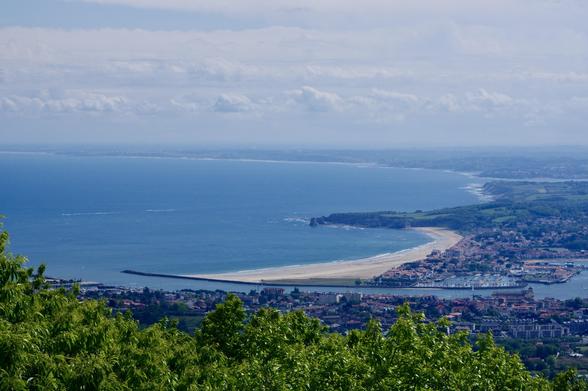A panoramic view of a coastline featuring sandy beaches, a harbor, and lush greenery in the foreground. The sea is calm with distant hills visible under a cloudy sky.