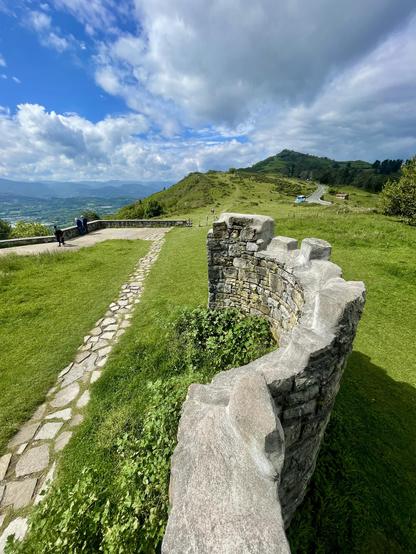 A scenic view featuring a stone wall in the foreground, set against rolling green hills under a partly cloudy sky. A winding stone path leads through the landscape, with a few people walking in the distance.