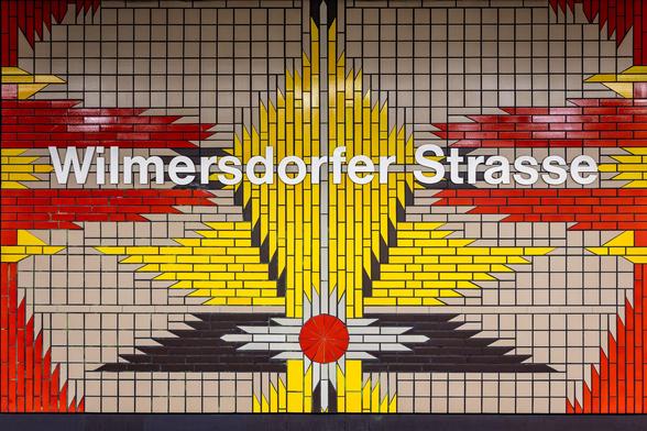 Black, red, yellow and white mosaic tiles representing a flower at Wilmersdorfer Strasse U-Bahn station in Berlin.  The pattern of the tiles are meant to represent a lily which can be found on the crest of Wilmersdorf. Wilmersdorfer Strasse U-Bahn station was opened in 1978 and designed by Rümmler.