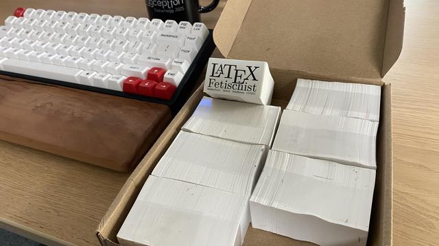 A box with a lot of stickers saying “LaTeX Fetischist” with the subtitle “Underful \hbox (badness 10000)”.

In the background is a keyboard with a wrist rest and a cup on the same desk.