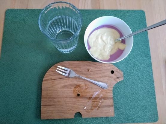 Auf einem Holztisch liegt ein rechteckiges, grünes Tischset. Darauf ein Holzbrett in Form eines Bäres, darauf eine Kuchengabel neben Brotkrümeln. Darüber rechts eine weiße Porzellanschale mit fliederfarbenem Joghurt und Vanillepudding oben drauf.
Links ein geriffeltes Wasserglas zu 2/3 gefüllt.