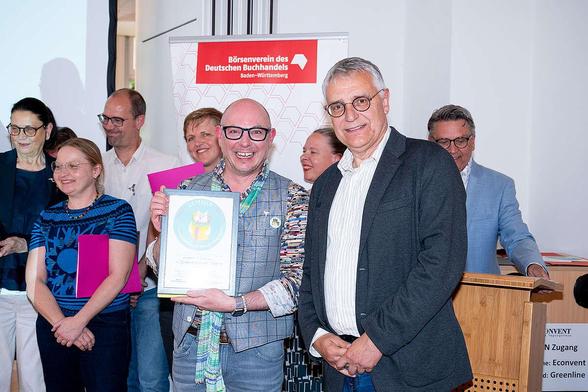 Buchhändler Markus Schneider mit seiner Urkunde daneben Staatssekretär Arne Braun; im Hintergrund weitere Preisträger.