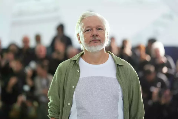 Photo of Julian Assange, looking quite well and wearing a white t-shirt with the names of the Palestinian children on the front.