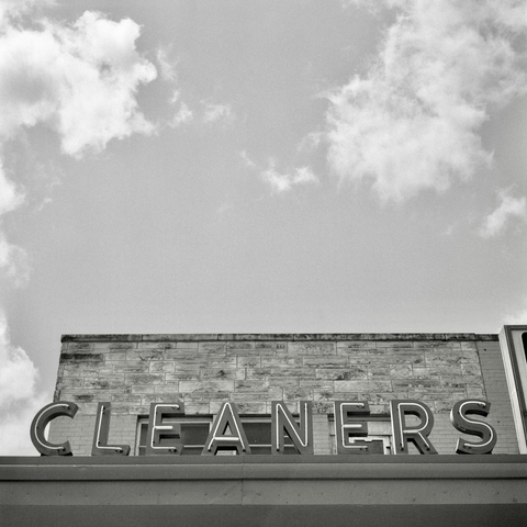 A black and white film photo. A vintage neon sign CLEANERS stands in front of a stone facade and a great big cloudy sky in Austin, Texas. Tuesday, April 22, 2025.