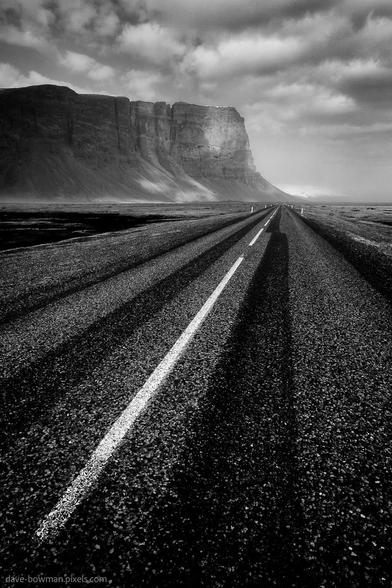 A monochrome photograph of a quiet, deserted road cutting through the stark landscape of southern Iceland. The white lines along the tarmac lead the eye toward a dark, looming cliff face in the distance. Heavy clouds hang low in the sky, adding a sense of weight and emphasising the isolation and scale of the open surroundings.