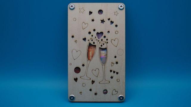 Verschraubte Holzkarte mit ausgeslasertem Motiv zweier Sektgläser, dekoriert mit Herzen und Sternen, hinterlegt mit Geldscheinen als romantisches Geschenk.