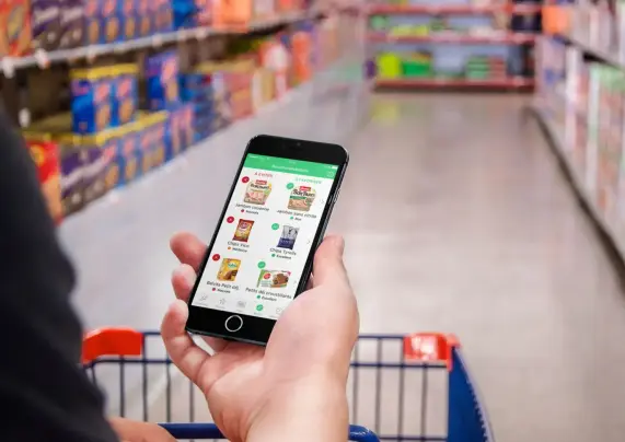 Application Yuka sur un téléphone mobile dans le main d'un consommateur poussant un caddie bleu dans les rayons d'un supermarché