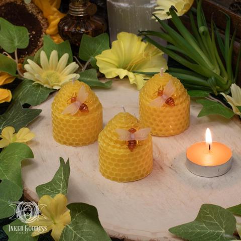 Three examples of the "Beehive Beeswax Candle" are arranged on a raw wood slice. They are made of rolled beeswax and are in the shape cylinders topped with cones. A small plastic bee is pinned into each candle. The candle is about votive candle-sized. Presented by Inked Goddess Creations.