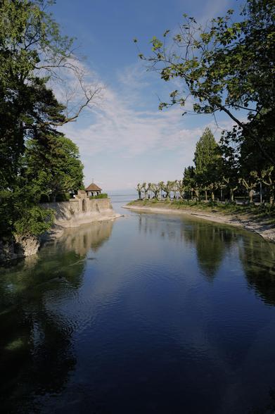 Der Inselkanal Konstanz, gesäumt von Bäumen, mit einer kleinen Steinkonstruktion und einer Hütte mit Spitzdach links. Im Wasser spiegelt sich der blaue Himmel. Getrimmte Bäume säumen das rechte Ufer.