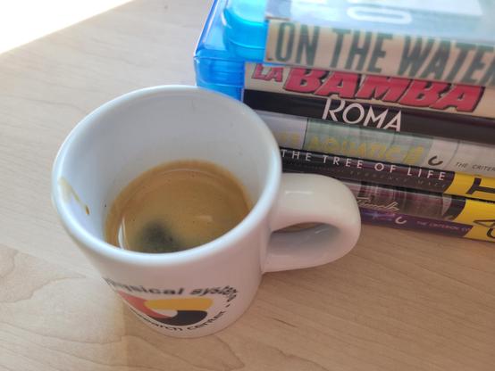 A white, 3 ounce, ceramic espresso (demitasse) cup with a shot of espresso, on a birch desk. The blue text on the cup reads "Cyber-Physical Systems Research Center" and wraps in a circle around the logo, which looks like a loosely interlaced, rounded triquetra with thick knots in solid red, yellow, and blue (each its own color). 

Behind it at right is a stack of 6 Blu-ray Criterion Collection movies checked-out from the public library: 1) La Bamba; 2) Roma (2018); 3) The Life Aquatic; 4) The Tree of Life; 5) The Asphalt Jungle; and 6) Gummo.