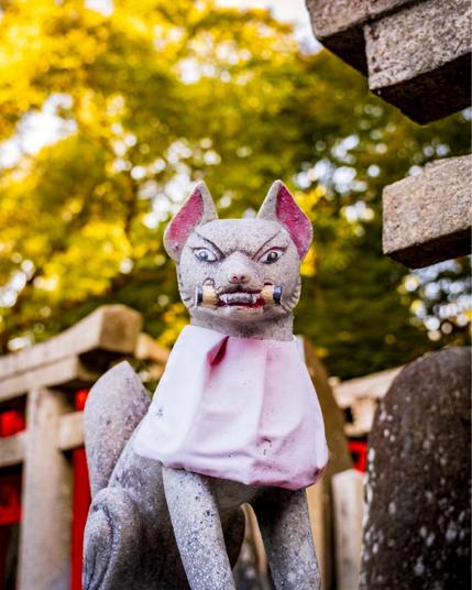 A stone fox statue at Fushimi Inari Shrine in Kyoto, Japan. The fox, known as a kitsune, has a stern expression, holds a key in its mouth, and wears a slightly weathered pink bib. The background features traditional stone structures and lush green trees illuminated by soft, golden light.