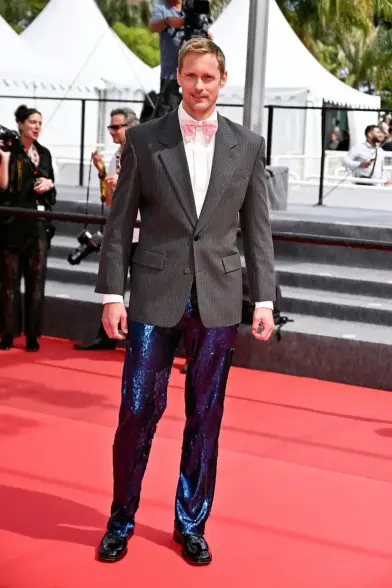 Alexander Skarsgård on the red carpet in Cannes. He wears a white shirt, with a grey pin stripe suit jacket, a light pink bow tie, blue rhinestone trousers and black shoes. It's a totally messy clash of styles, colors and textures.