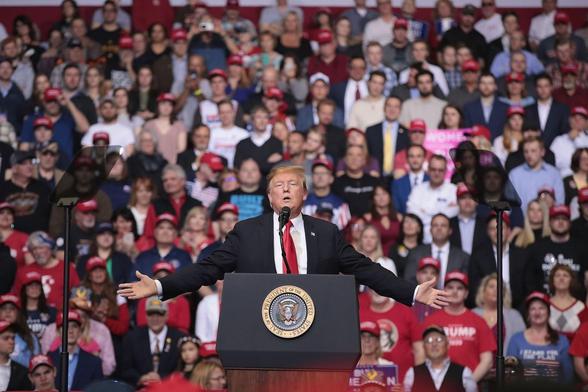 A color photo of a Trump rally. President Trump is in the foreground, behind a podium with his arms outstretched, eyes closed, in mid-sentence. Behind him is a large crowd.