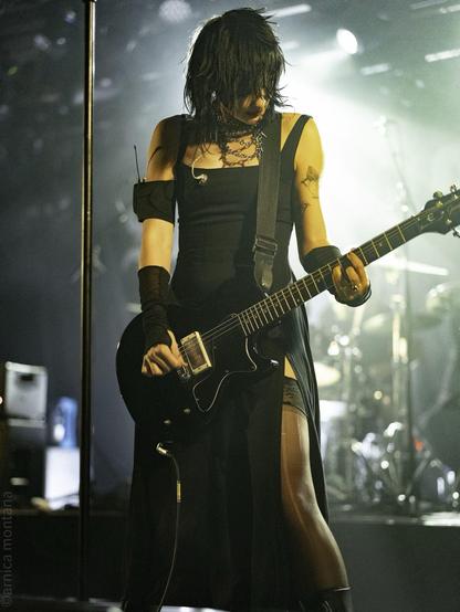 A musician performs on stage, holding an electric guitar while dressed in a black outfit. The scene is illuminated by stage lights, showcasing a vibrant atmosphere. The musician has long, dark hair and visible tattoos, contributing to a striking visual presence.