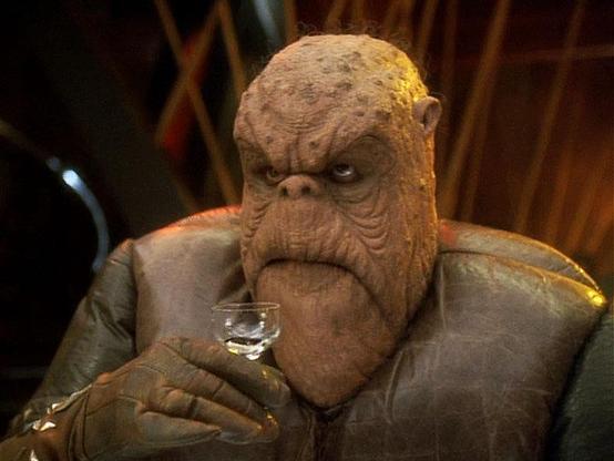 Star Trek Deep Space Nine scene. We're looking at an alien named Morn. Morn is looking up while having a sit and holding up a wee little cocktail glass. Morn is wearing a soft padded jacket that looks like one of those nice old fancy leather couches with some weathering. His face is thumb shaped, w/tiny lil ears and a smaller and higher than normal little nose. Skin sort of looks like droopy bark on a tree? This probably makes no sense, Morn is tricky to describe because he contains so many multitudes. No caption. 